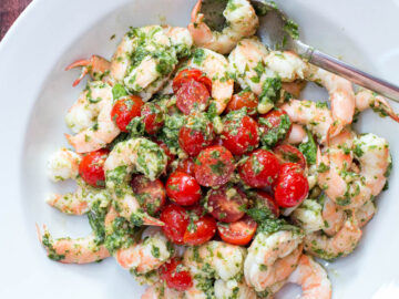 Cooked shrimp in a deep plate with halved cherry tomatoes and basil pesto. Garnished with Pine nuts.