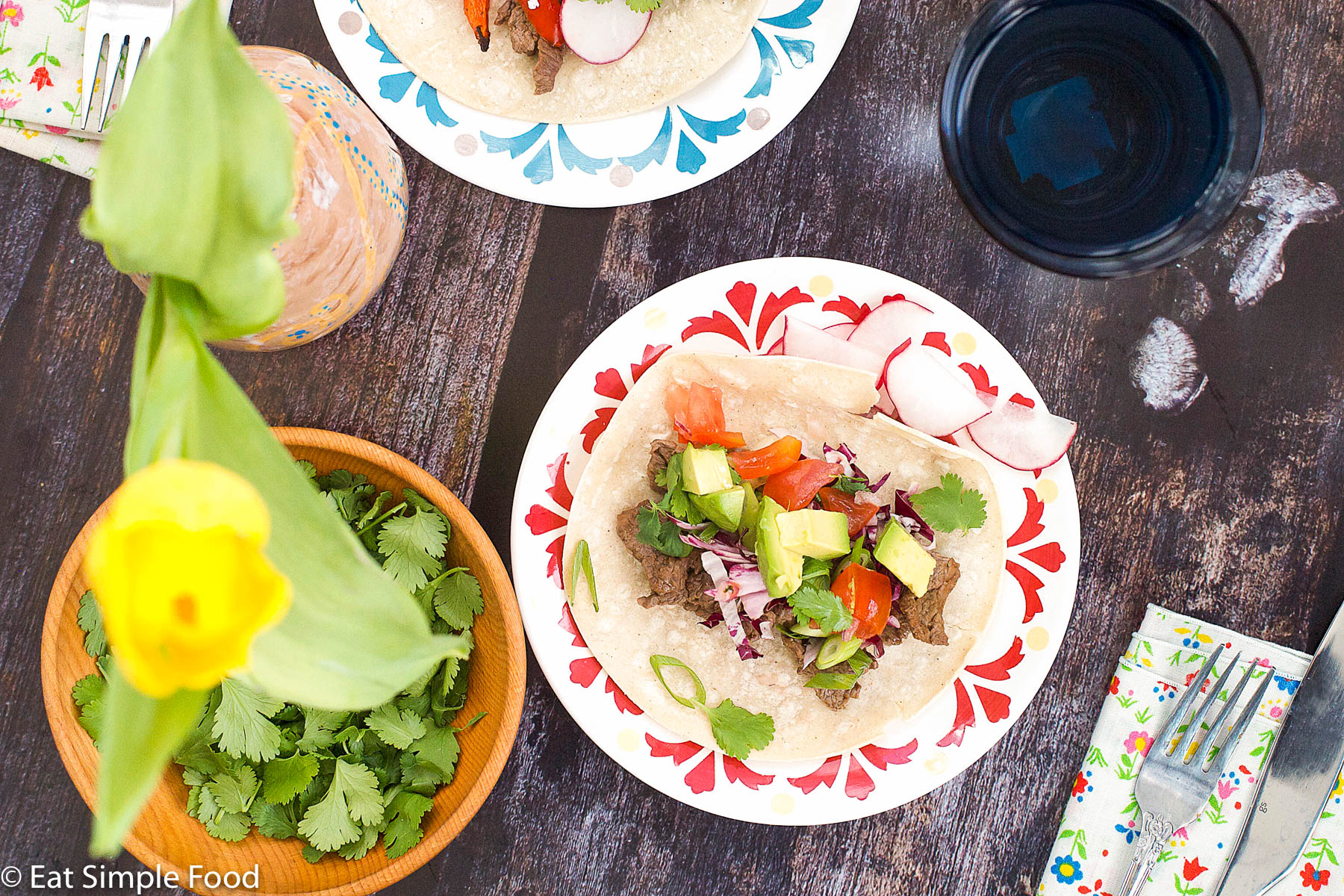 2 Plates with one taco each. Flour tortilla filled with sliced cooked steak, tomato salsa, red cabbage, tomatoes, and cilantro. sliced radishes on the side. Glass of water, fork, knife and napkin. Top view.