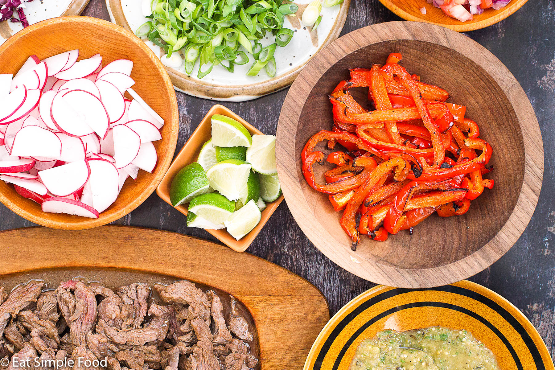 Taco Ingredients. Top view. Bowls and plates filled with: sliced radishes, roasted sliced red peppers, lime wedges, green salsa, sliced green onions, cooked sliced steak.