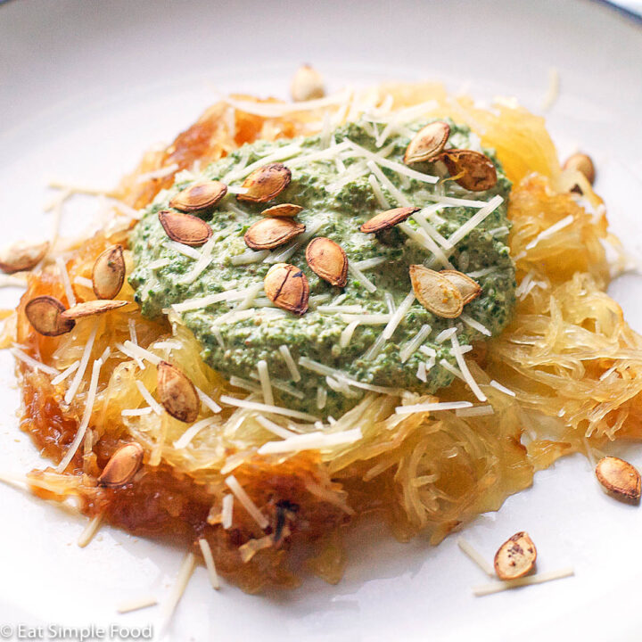 Yellow Spaghetti Squash "pasta" with green kale and parmesan pesto with toasted squash seeds on top. Ona white plate with black rim.