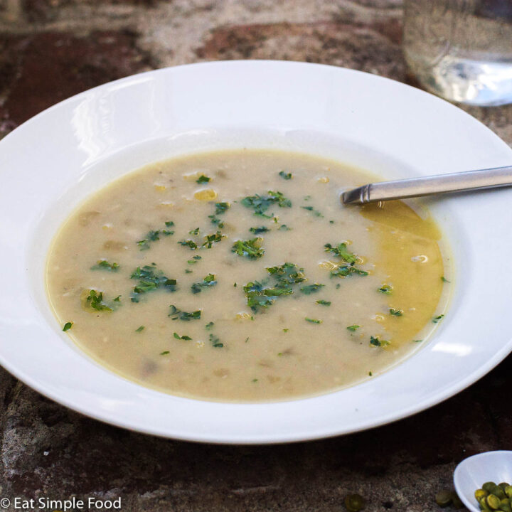 White shallow bowl of green/brown split pea soup with green parsley garnish. Spoon in the soup.