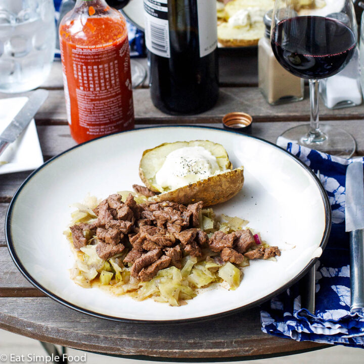 One plate of browned strips of beef with cooked cabbage and a baked potato with sour cream. Bottle of red wine, 2 full glasses and a bottle of sriracha hot sauce.