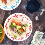 2 Plates with one taco each. Flour tortilla filled with sliced cooked steak, tomato salsa, red cabbage, tomatoes, and cilantro. sliced radishes on the side. Glass of water, fork, knife and napkin. Top view.