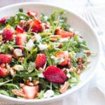 quartered strawberries, chopped pecans, and crumbled goat cheese on arugula salad on a shallow bowl/plate.