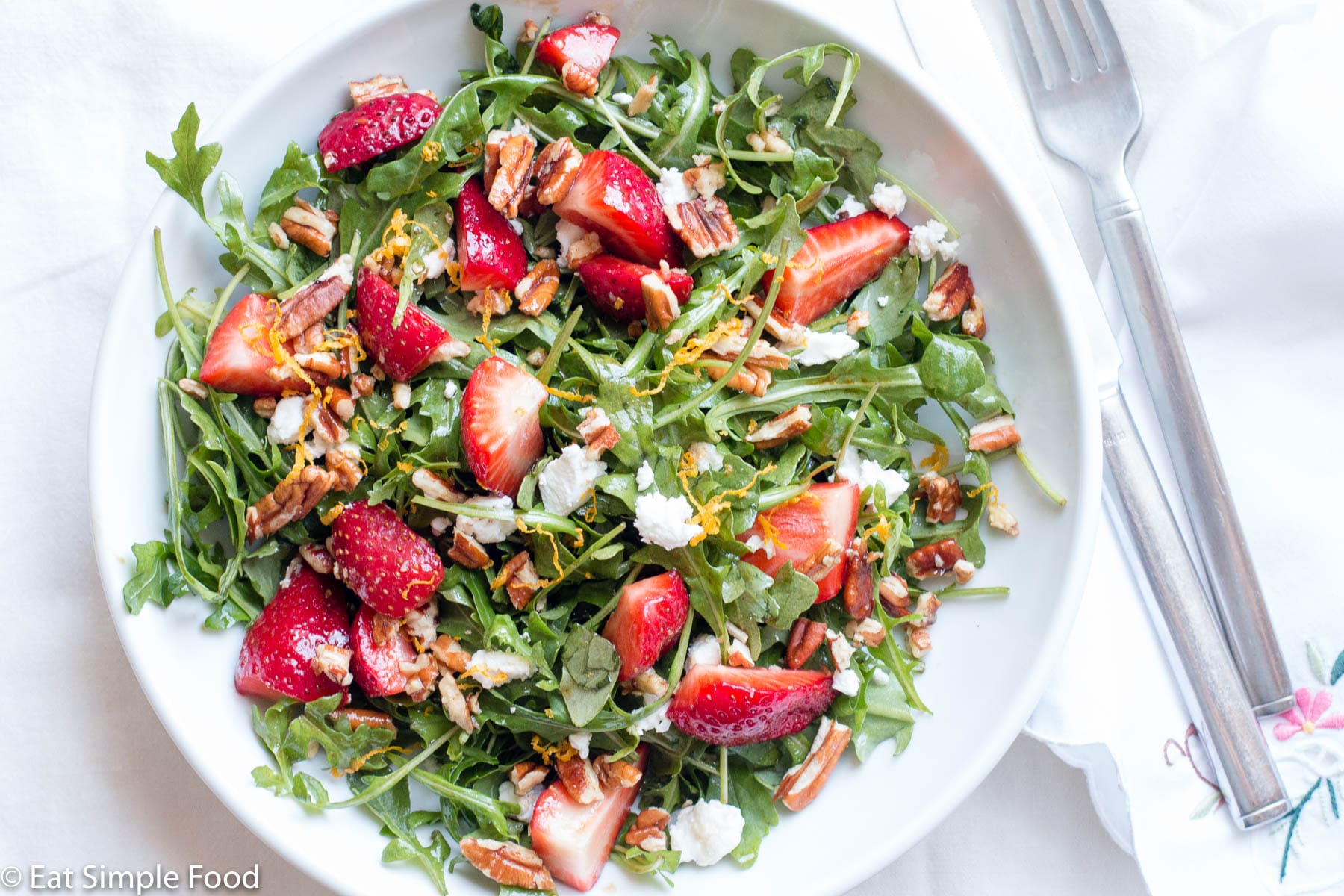 quartered strawberries, chopped pecans, and crumbled goat cheese on arugula salad on a shallow bowl/plate. top view.