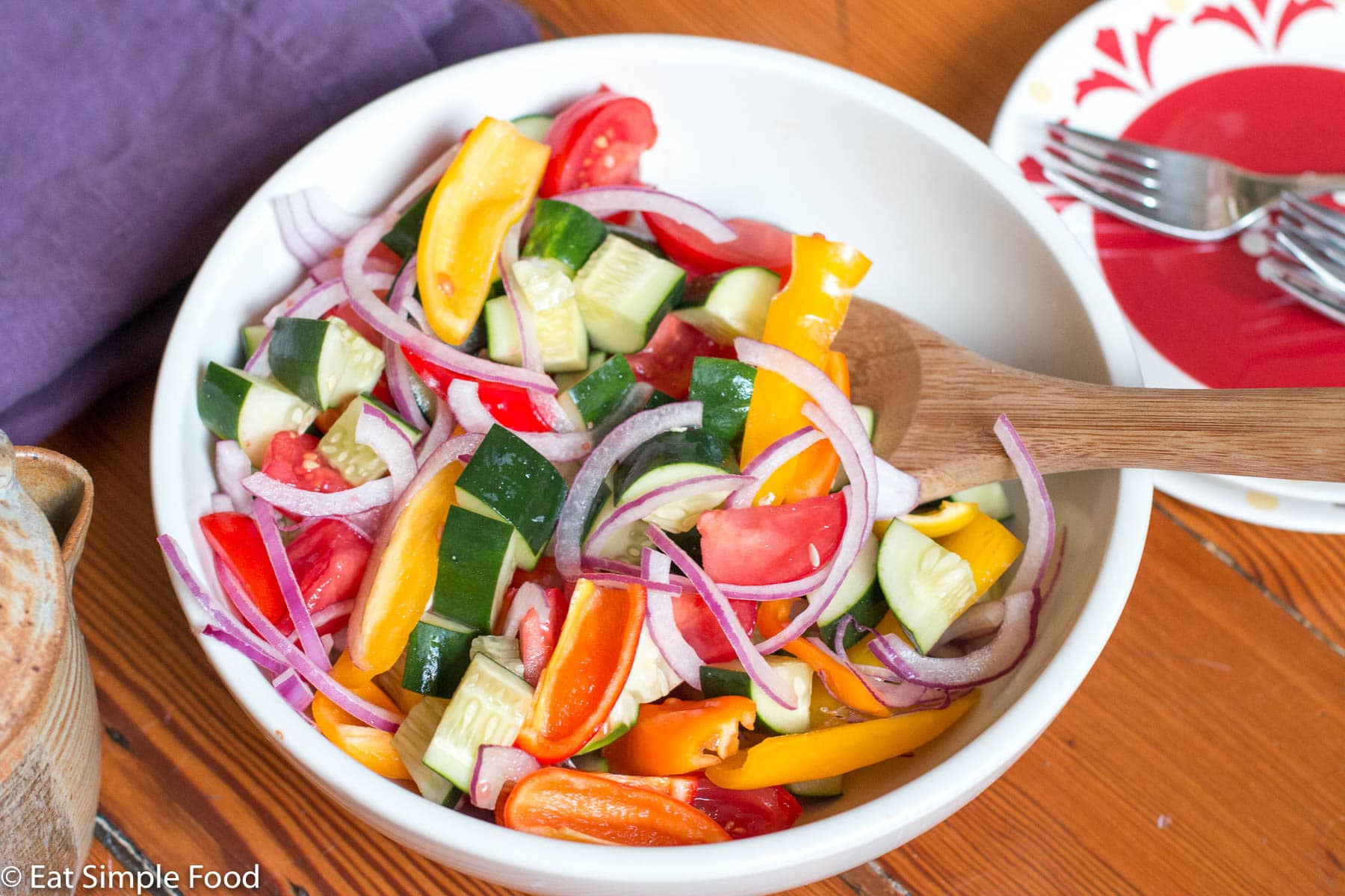 White bowl of chunky red onions, cucumbers, tomatoes, yellow and red peppers salad. Wood spoon it it. side view.