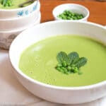 Large white bowl of green creamy soup garnished with fresh peas and mint leaves. Small bowl of peas and 3 bowls in background.