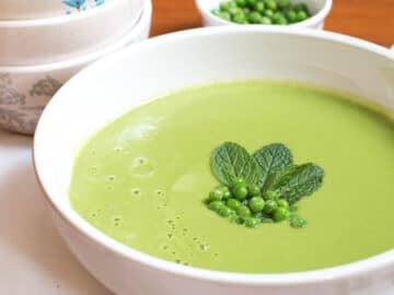 Large white bowl of green creamy soup garnished with fresh peas and mint leaves. Small bowl of peas and 3 bowls in background.