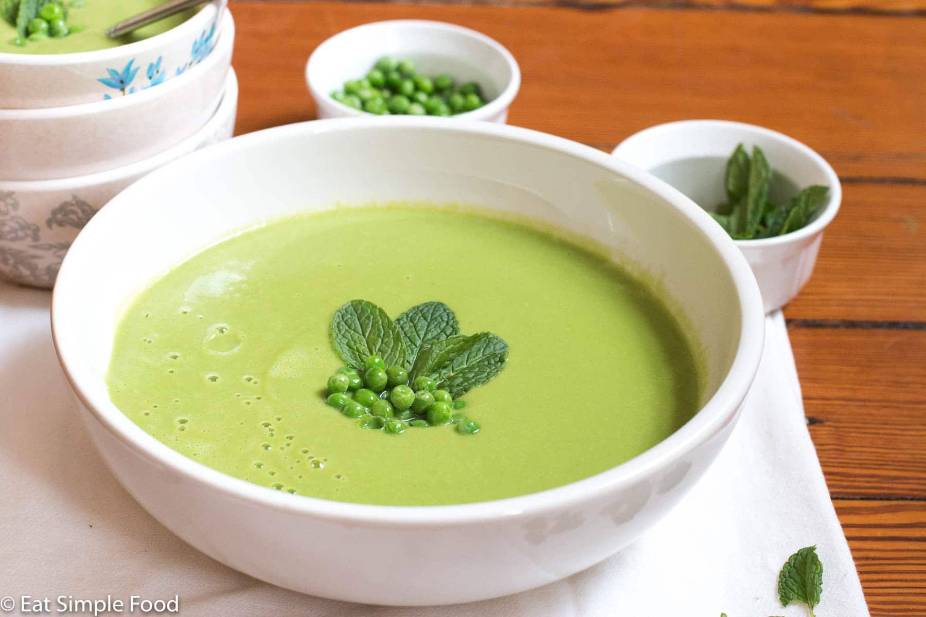 Large white bowl of green creamy soup garnished with fresh peas and mint leaves. Small bowl of peas and 3 bowls in background.