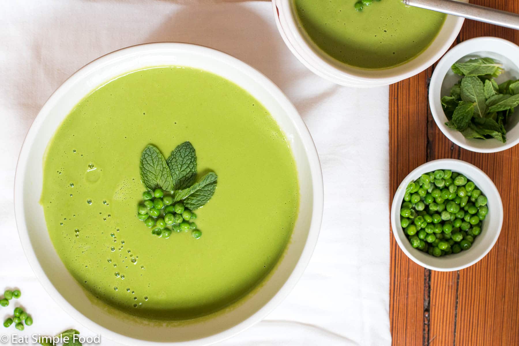Large white bowl of green creamy soup garnished with fresh peas and mint leaves. Small bowl of peas and bowl of mint leaves. Top view.