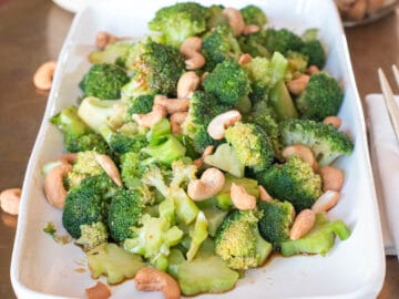 Broccoli w/ Tamari (Soy Sauce) & Cashews on a rectangle white plate on a copper table. Cashews in jar in background.