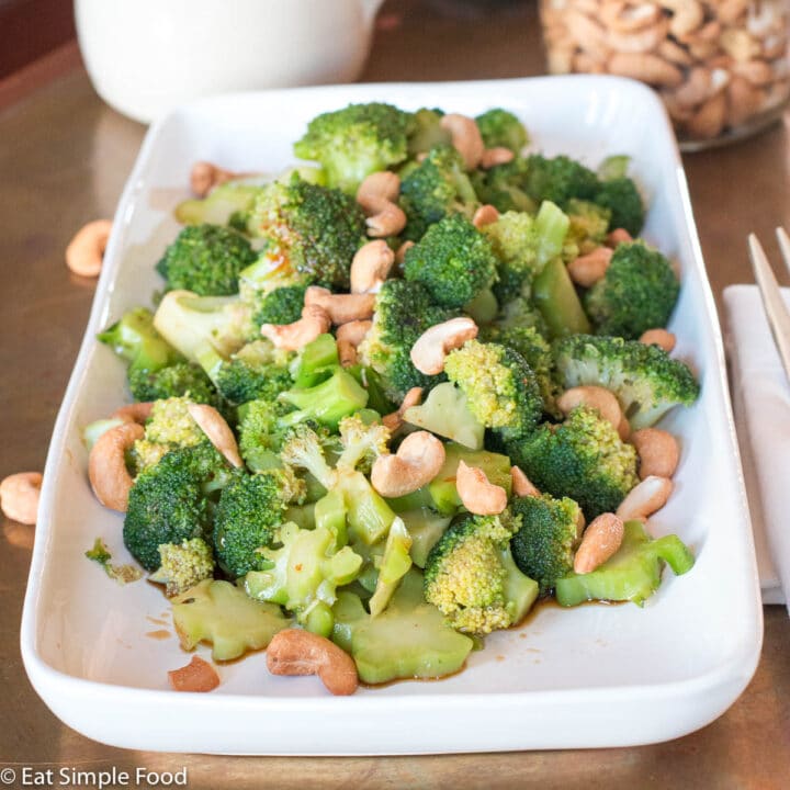 Broccoli w/ Tamari (Soy Sauce) & Cashews on a rectangle white plate on a copper table. Cashews in jar in background.