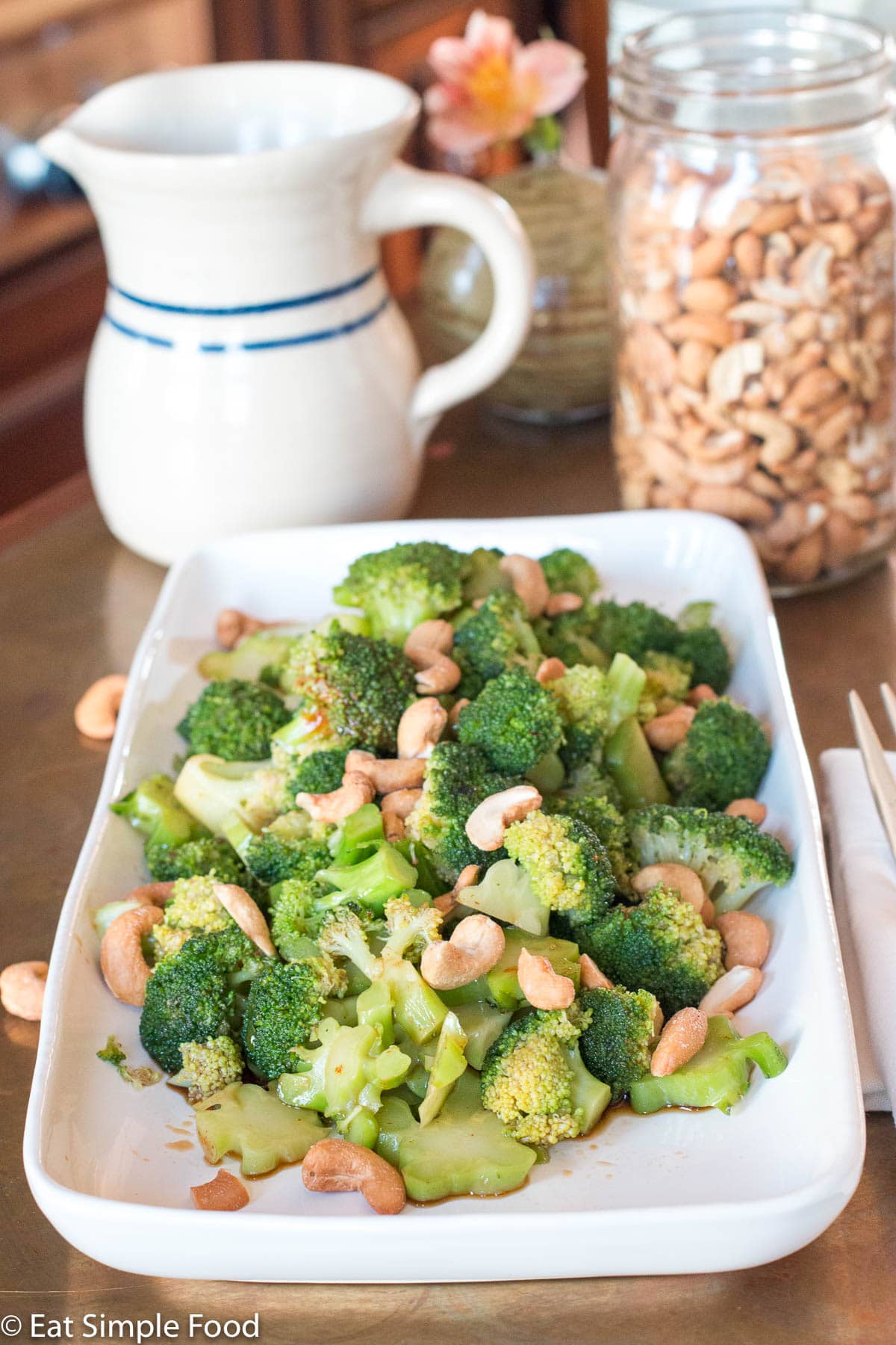 Broccoli w/ Tamari (Soy Sauce) & Cashews on a rectangle white plate on a copper table. Cashews in jar in background.