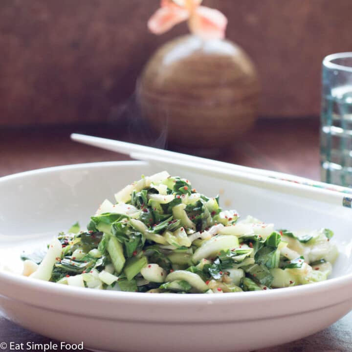Sliced bok choy in a shallow bowl with white chopsticks sitting on the side. Glass of water in the background