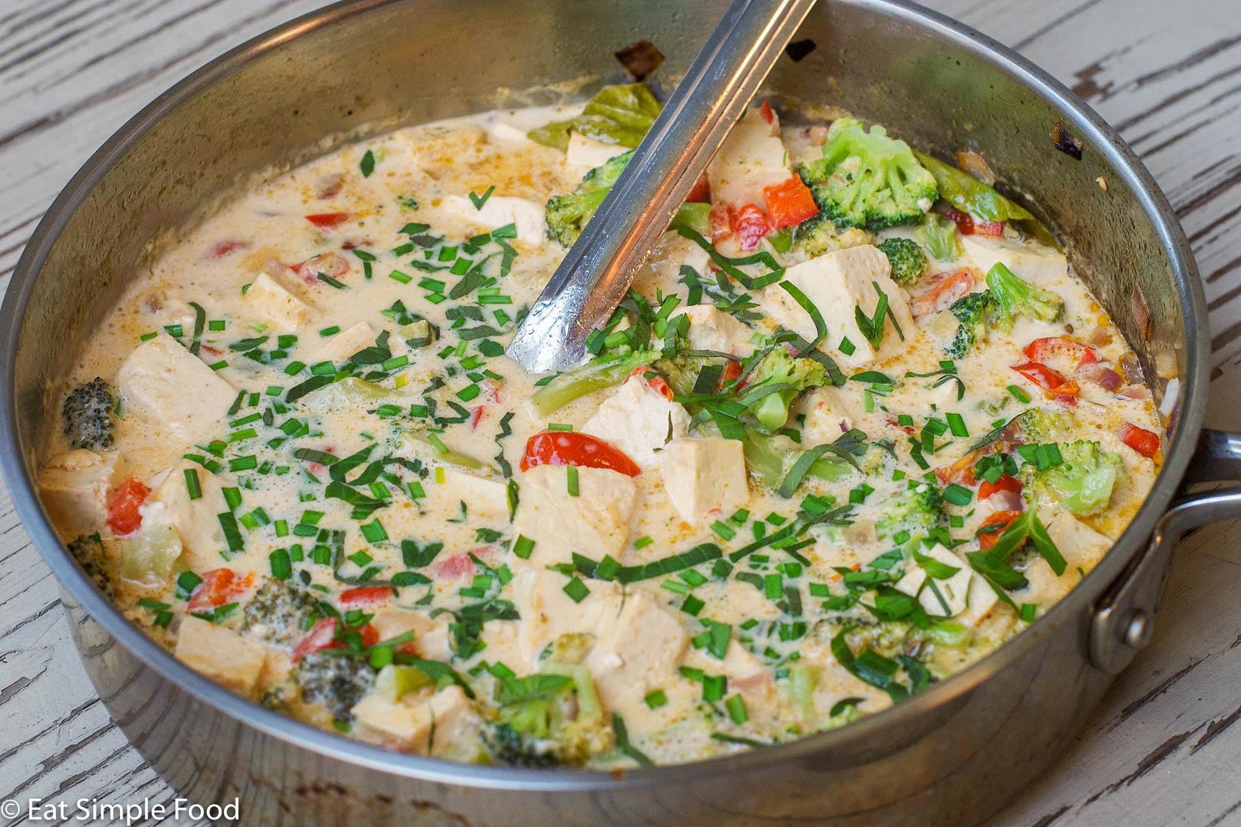 Deep Stainless steel pan filled with cubed tofu, chunked red peppers and broccoli and garnished with sliced chives and basil. Serving spoon hanging out the side. close up.