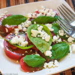 Slices of Tomato, basil leaves, crumbled goat cheese on a white square plate with two forks.