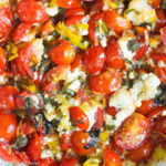 Close up top image of cooked cherry tomatoes, sliced basil, creamy chunks of goat cheese, sliced pepperoncini, and garlic in a bowl.