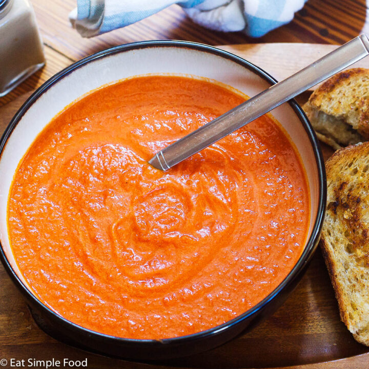 White bowl with black rim of red tomato bisque soup on a wood rectangle plate. Grilled cheese sandwich cut in half diagonally on the side.