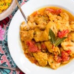 White bowl of chicken thighs in a yellow sauce with tomatoes, onions, and capers. Yellow Rice in bowl on the side. Top view.