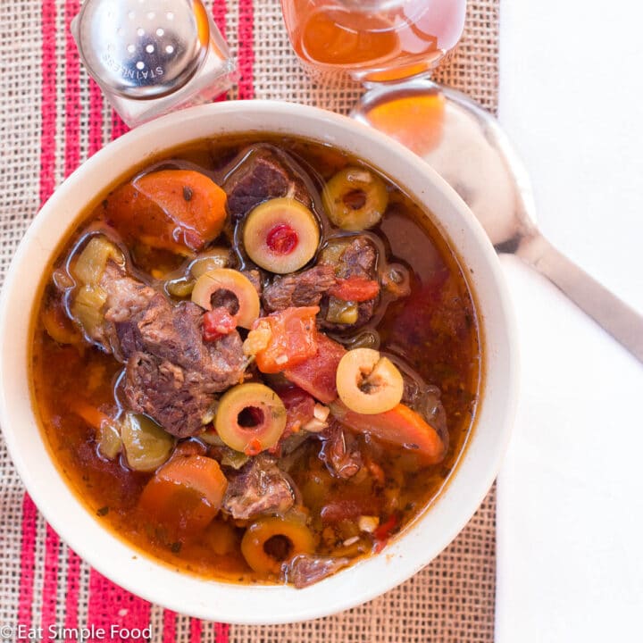Top view of bowl of beef stew with green olives, carrots, green chilis, garlic, and tomatoes. Salt shaker, spoon, and bottletop drink on the side.