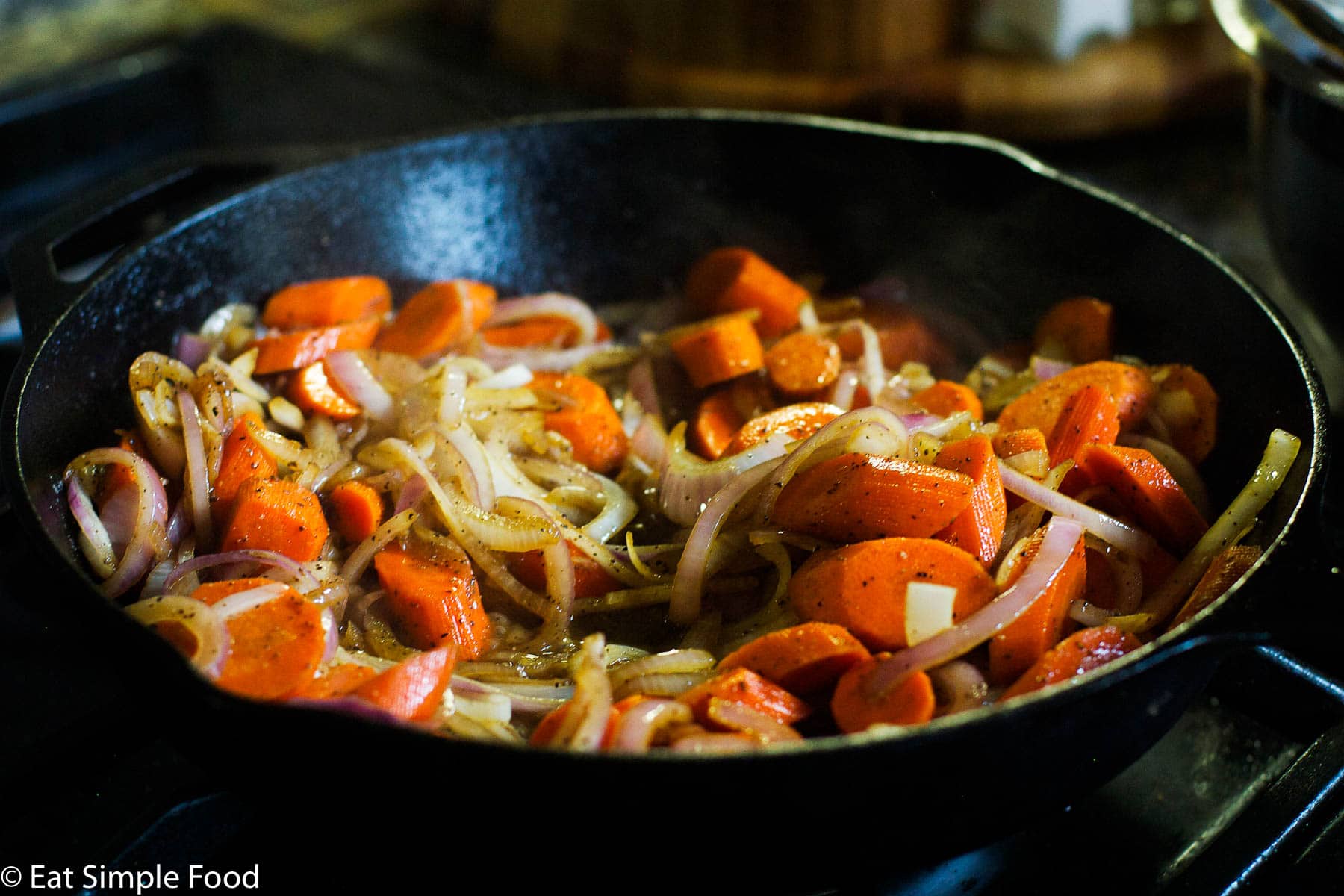 Chunked Carrots and Sliced Onions cooking in a large cast iron pan on a gas stovetop.
