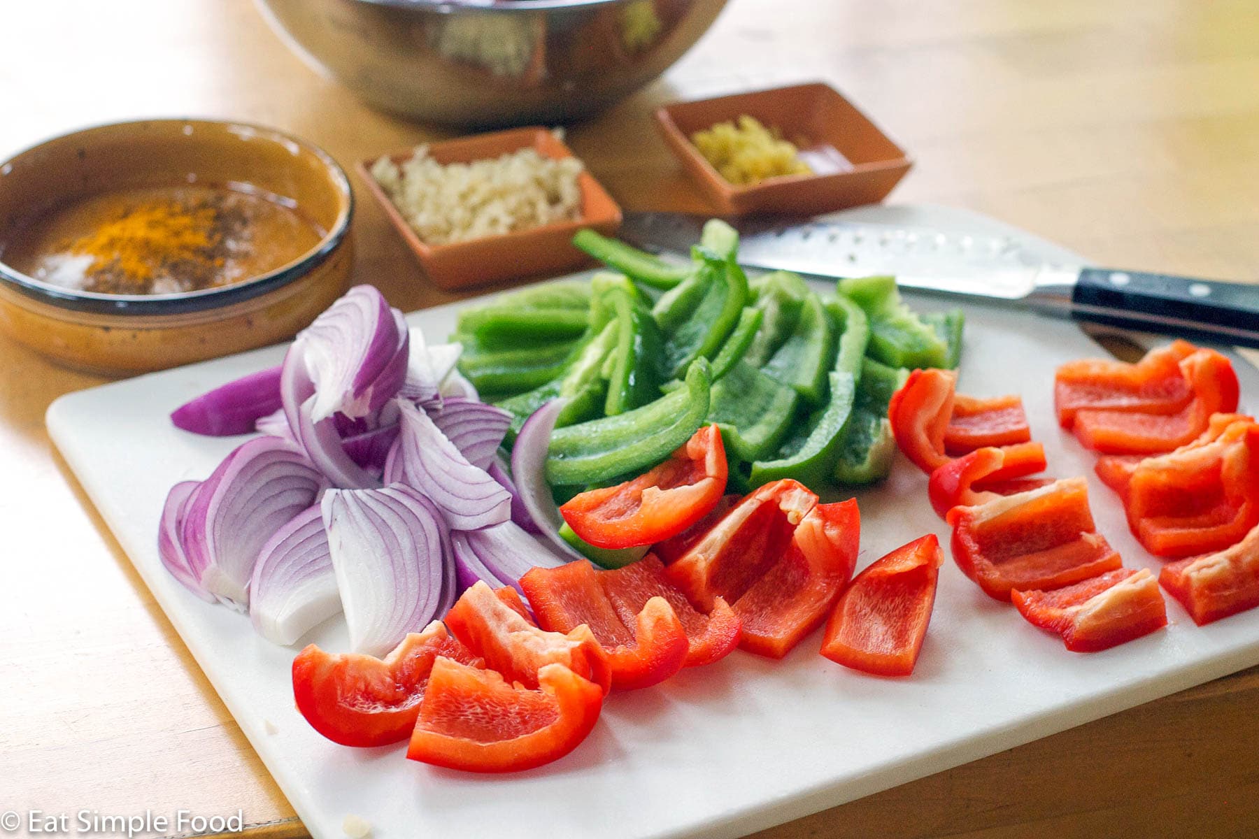 white cutting board with knife on it and sliced red and green bell peppers and red onion. Minced ginger and garic in small orange bowls in the background