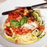Close up of Eggplant Parmesan with broiled on Mozzarella cheese and a basil garnish over zucchini noodles. White plate.