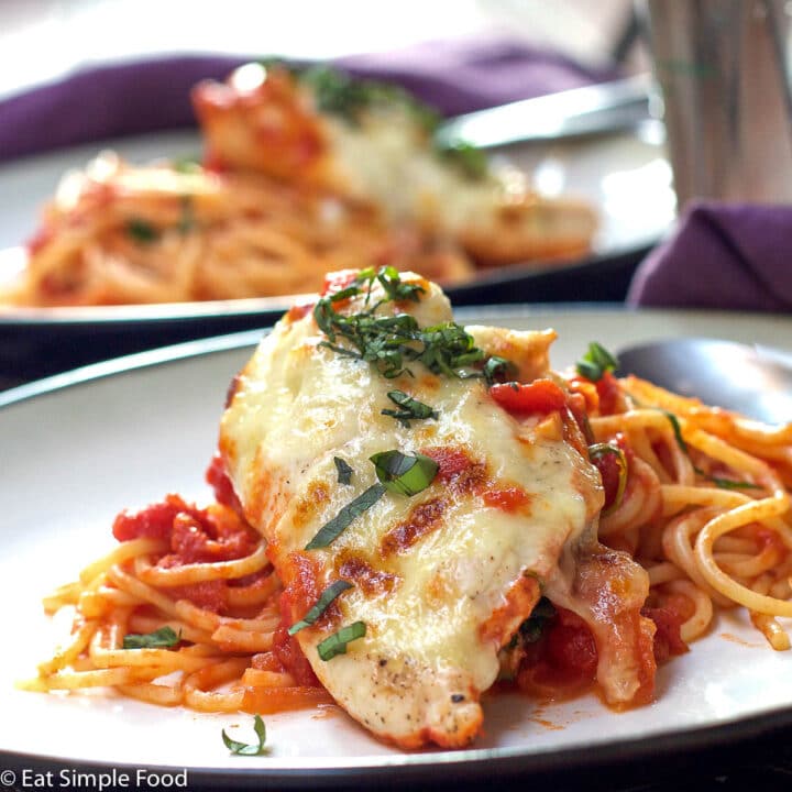 Chicken breast with Mozzerella broiled on over a bed of spaghetti with marinara. 2 white plates. Basil Garnish.