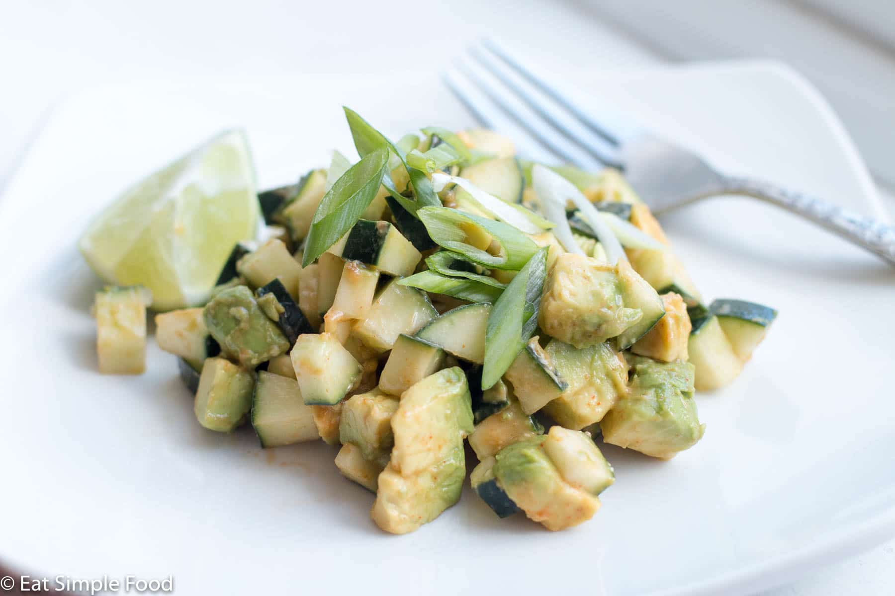 Small diced avocado and cucumber salad tossed in red hot sauce and garnished with green onions on a white plate. close up.