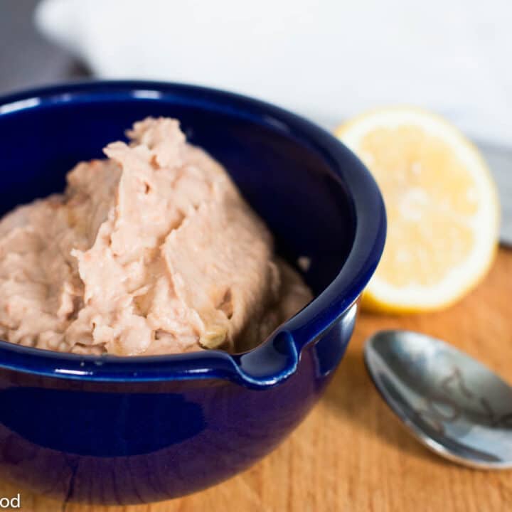 Blue bowl of blended tan pinto bean hummus on a wood cutting board with a lemon half and spoon in the background.