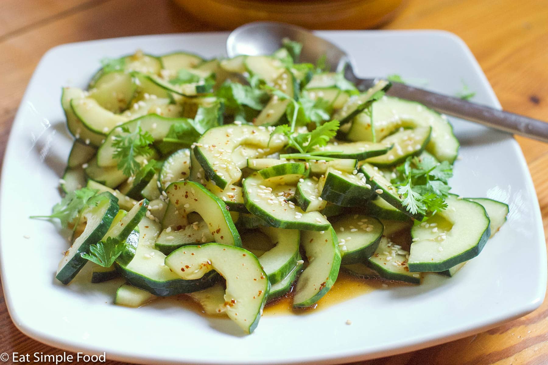Small white plate of halved cucumbers with seeds taken out tossed in a soy / sesame oil sauce and topped with sesame seeds and cilantro. Side view.
