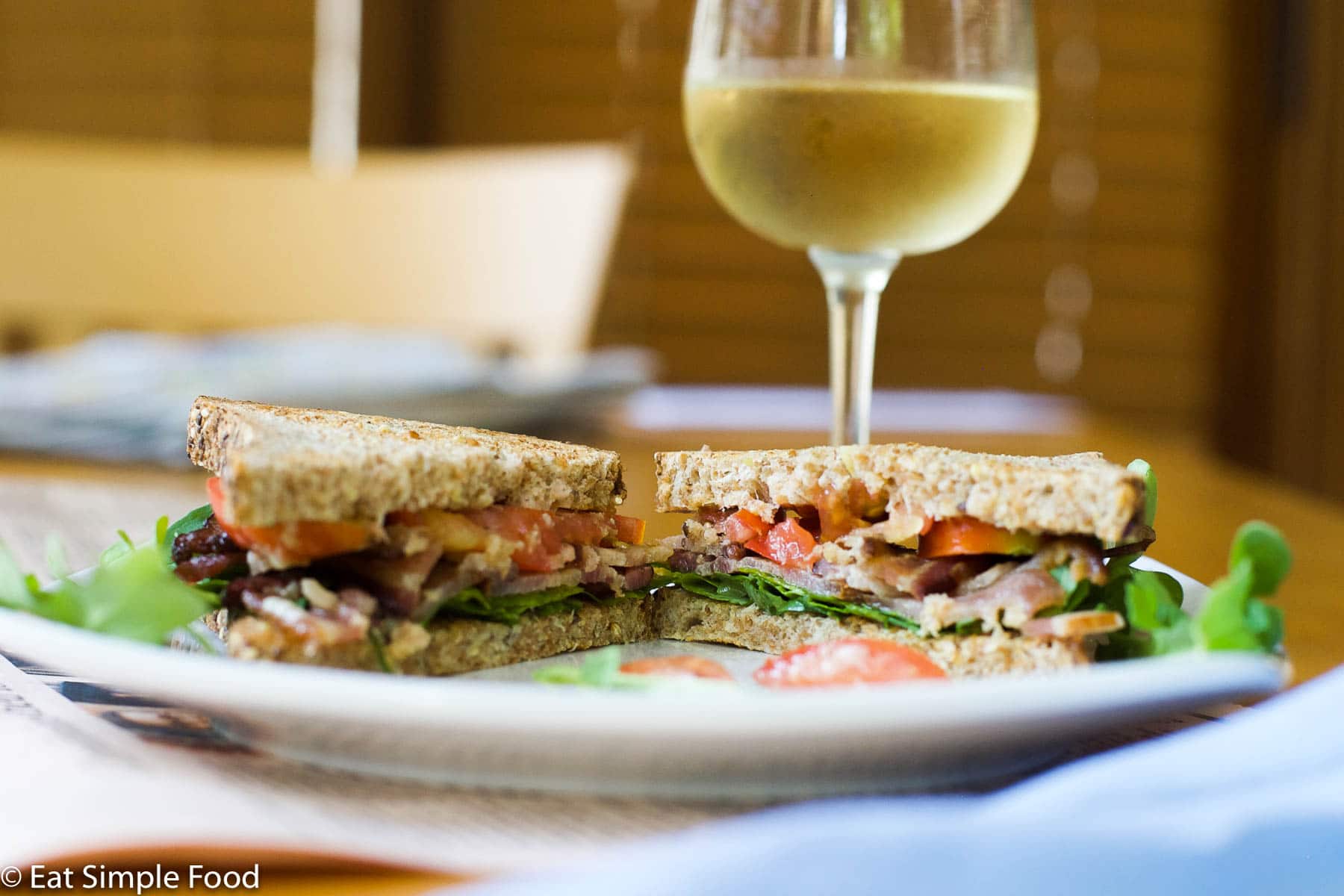 Bacon Arugula Tomato Stacked Sandwich on white plate. side view. Sliced in half. White wine in background.