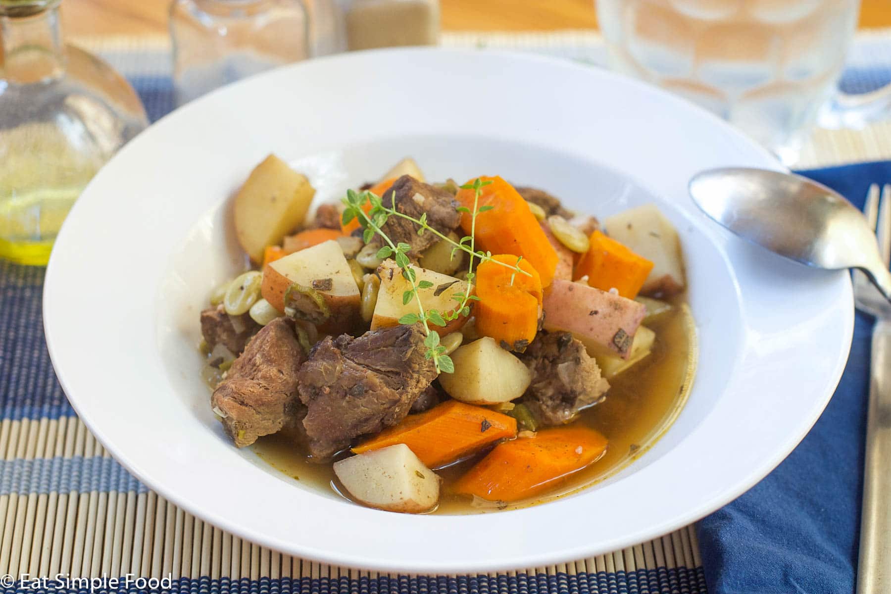 White shallow bowl filled with cooked browned beef, chunks of carrots and potatoes in a beef broth with thyme sprigs on top. Spoon and glass of white wine on side. Side view.