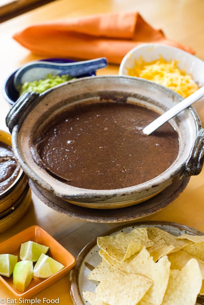 Pureed Black Bean Soup In a Brown Bowl (Side Angle) with A spoon with an orange container of lime wedges, bowl of tortilla chips, bowl of grated yellow cheese, and bowl of guacamole.