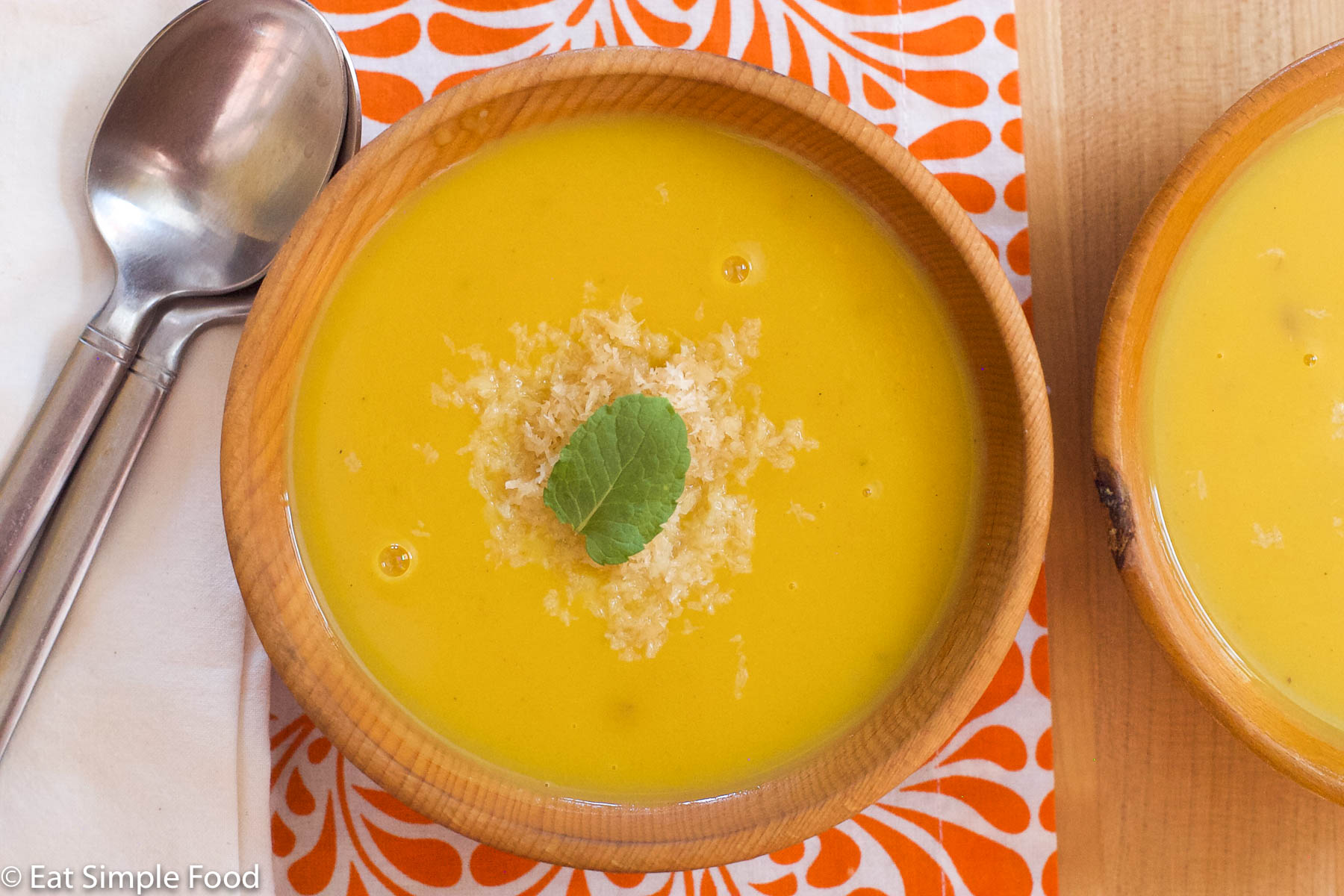 Wood bowl of pureed butternut squash soup with a garnish of toasted shredded coconut and a mint leaf. Spoons on the side. Top view.
