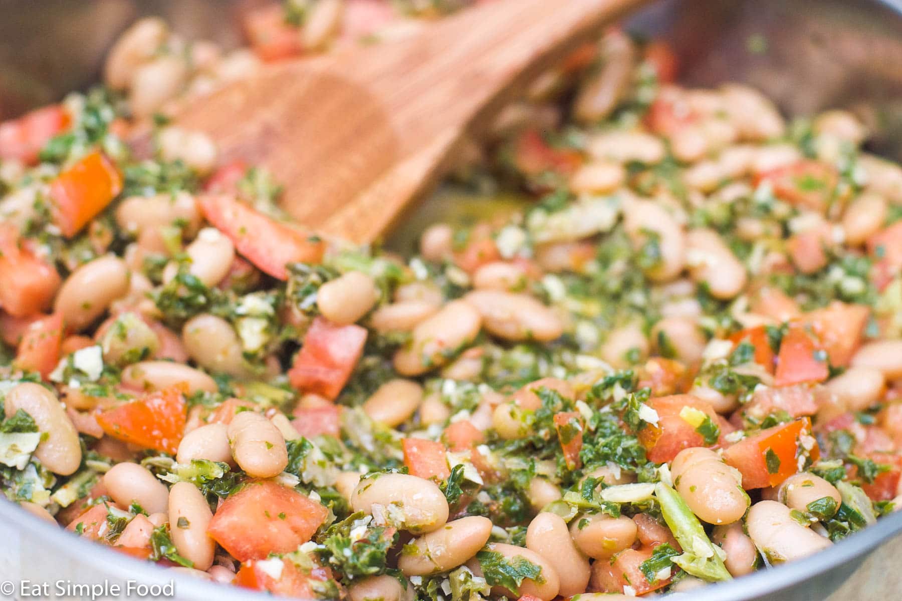 Stainless steel deep skillet filled with white beans and diced tomatoes with a lightly pulsed pesto of kale and almonds with a wood spoon. Close up.
