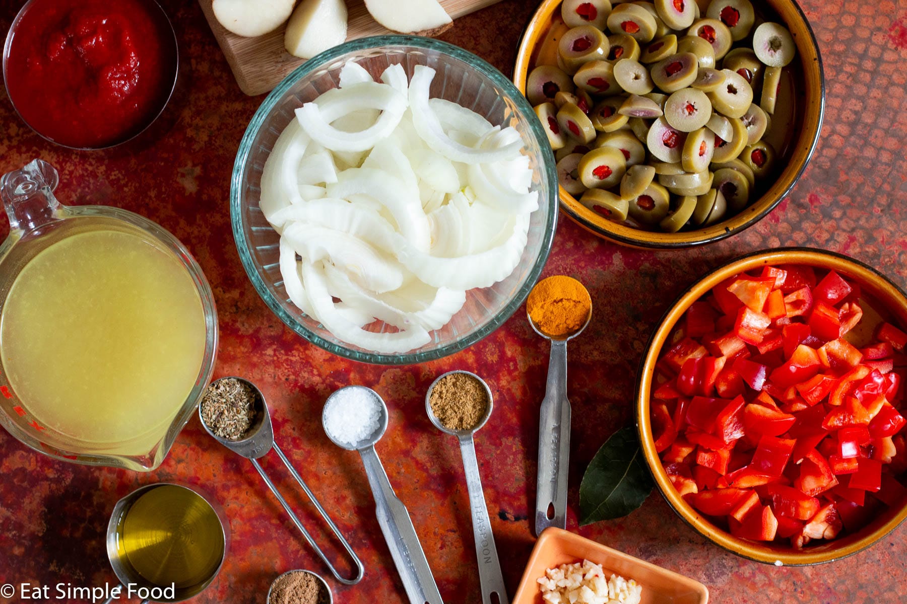 Sliced onions, diced red peppers, sliced green olives with pimentos, tomato sauce, chicken broth in different bowls with 5 teaspoons of colorful spices in teaspoons and a bay leaf on the table.