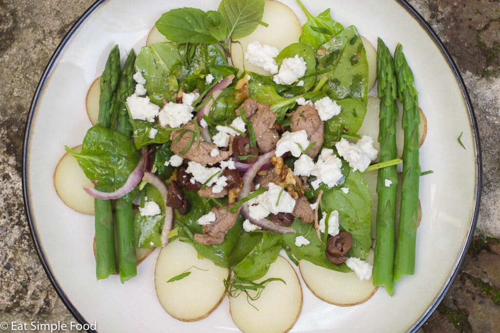 1 white plate with a layer of sliced cooked potatoes, blanched green asparagus spears, spinach, and seared lamb with crumbled feta garnish. Top view.