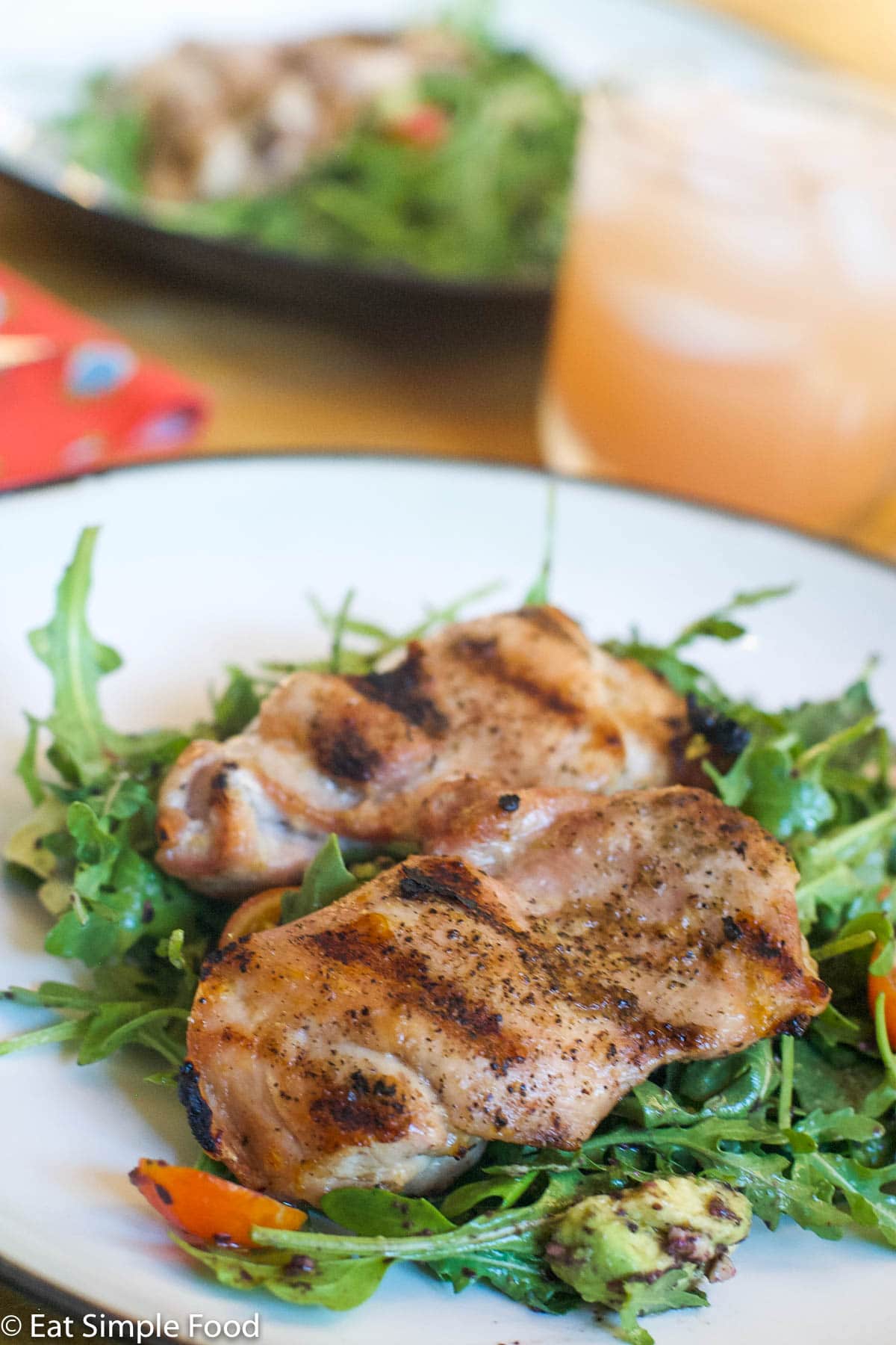 2 grilled chicken thighs over a bed of arugula salad w/ halved cherry tomatoes and diced avocado. On a white plate. Another plate in background with same thing.