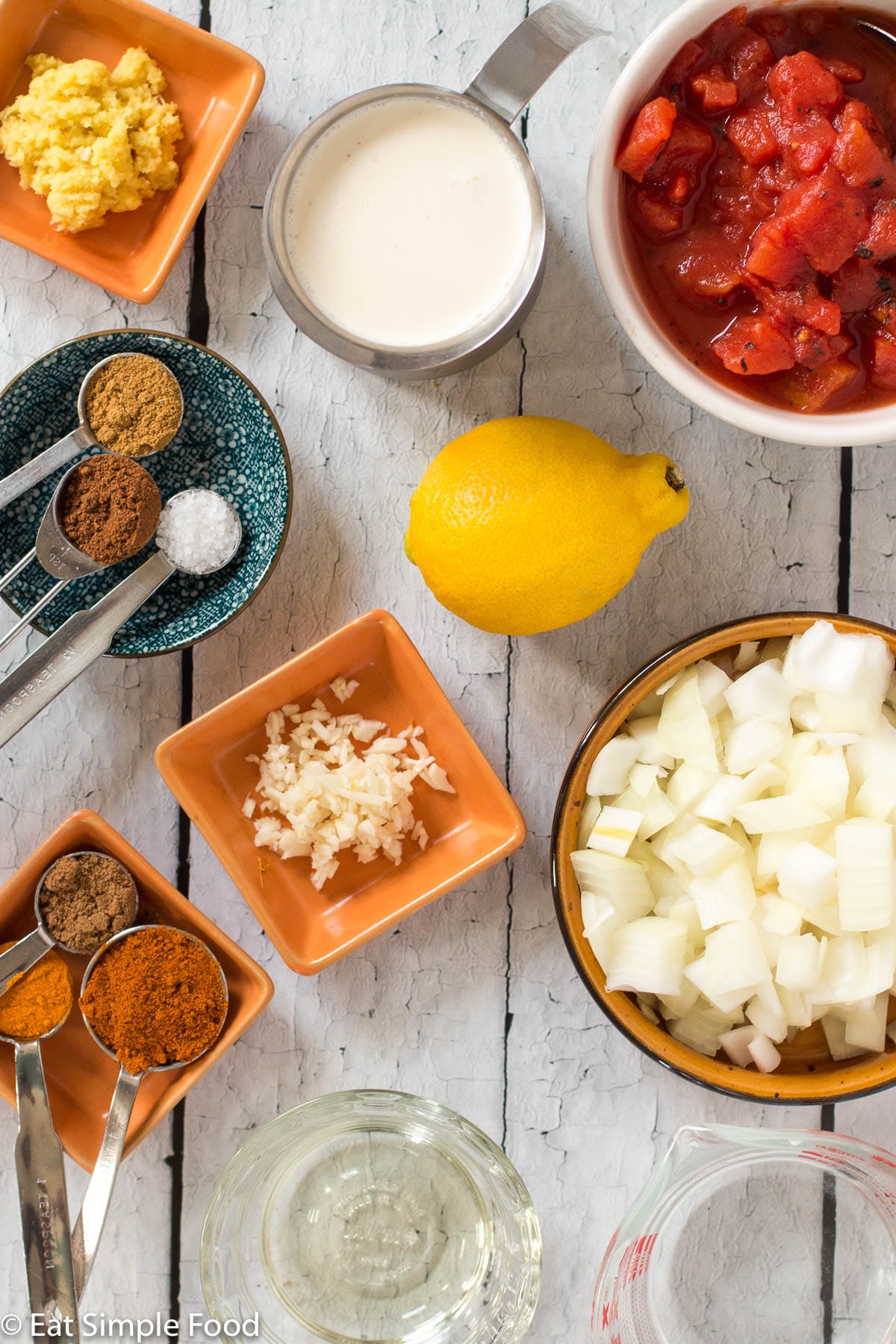 Ingredients in small cups and colorful containers: minced ginger, cream, diced tomatoes, garlic, 1 lemon, diced onions, vegetable oil, water, 6 teaspoons of colorful spices on white table.