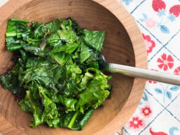 Wood bowl of Kale greens sauteed with a fork sticking out. white table. colorful flower napkin. top view.