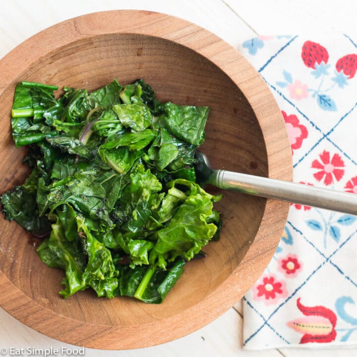 Wood bowl of Kale greens sauteed with a fork sticking out. white table. colorful flower napkin. top view.