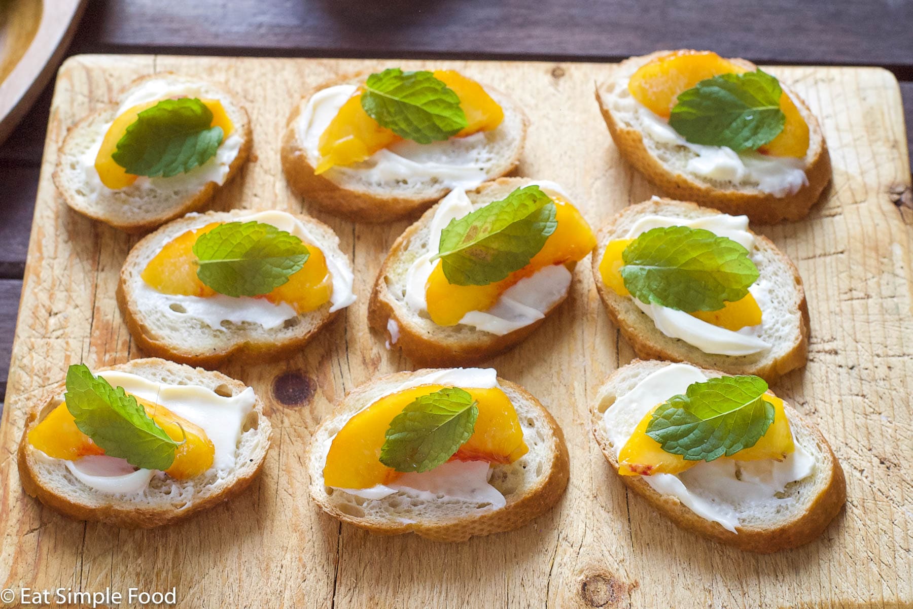 Crostini (little toasts) topped with spreaded mascarpone, a peach slice, and a mint leaf. Several crostini's on a wood cutting board. Top view.