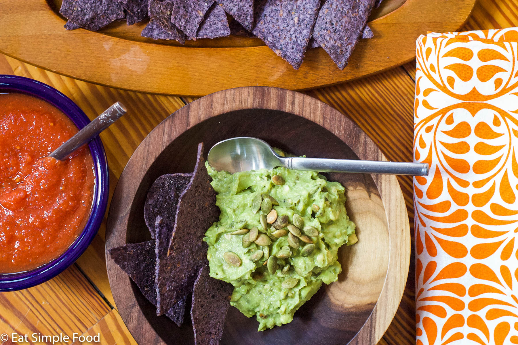 Guacamole in a wood bow with toasted pepita garnish with a spoon in bowl. Blue tortilla chips and bowl of salsa in background. Top view.