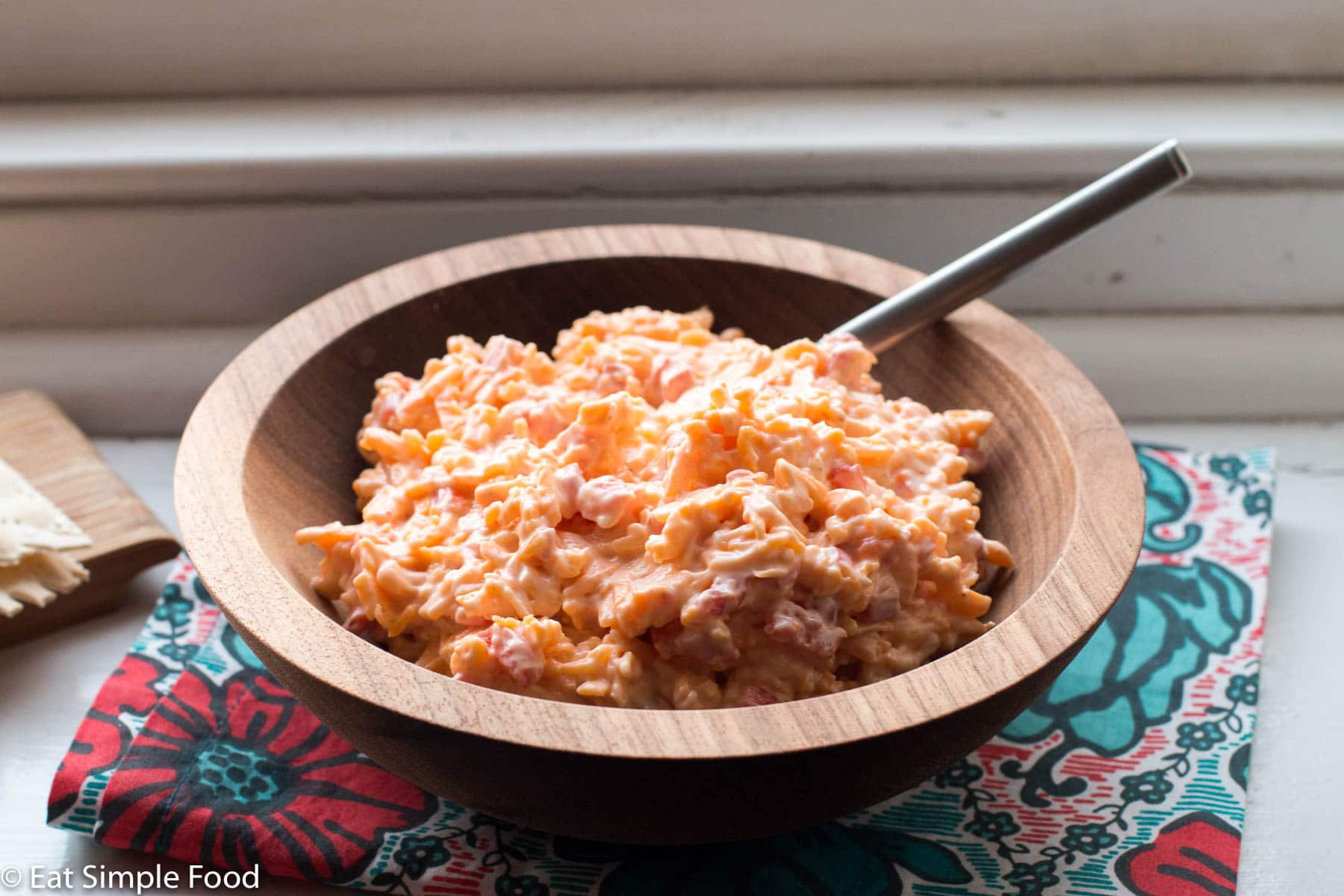Wood bowl of pimento cheese and a small spoon hanging out. Placed on a colorful napkin on a windowsill.