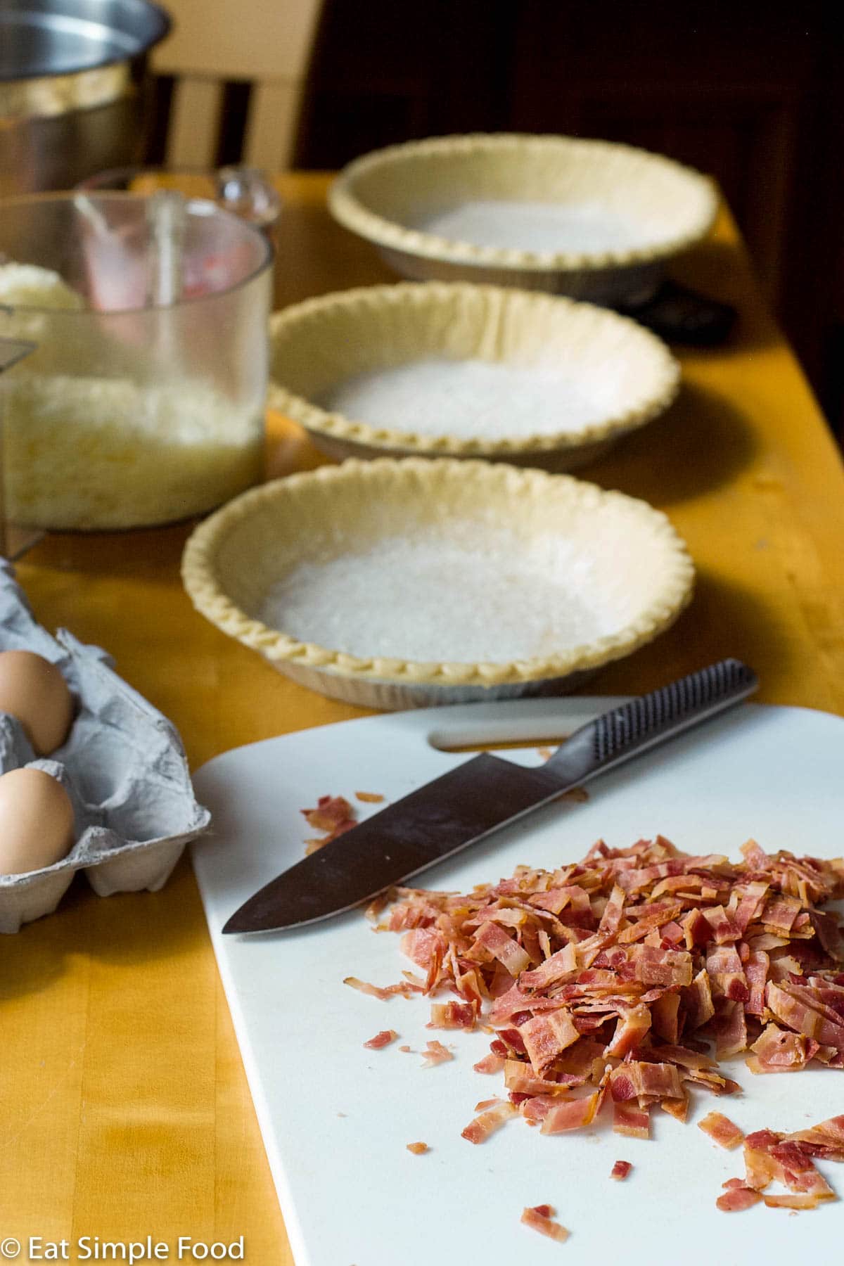 3 Quiche pie crust ready to be filled with eggs, cheese, and a cutting board of chopped cooked bacon.