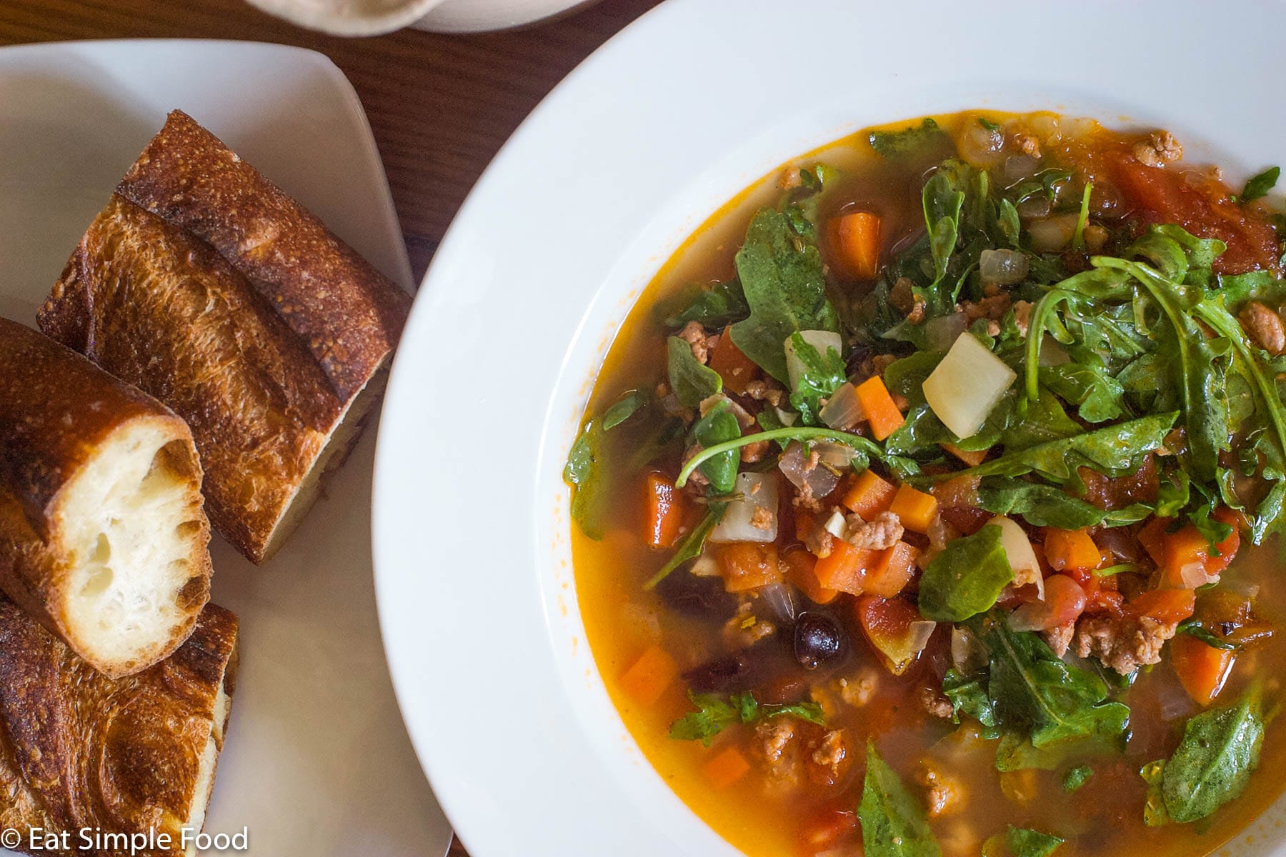 White bowl of tomato based soup with sausage, carrots, arugula, and white beans. Spoon and baguette on the side.