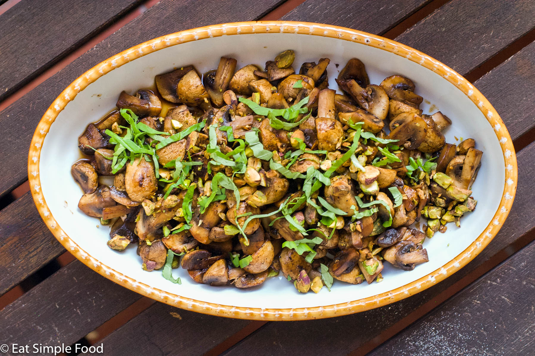 Cooked browned quartered mushrooms garnished with sliced basils and pistachios in a yellow baking dish. Top View.