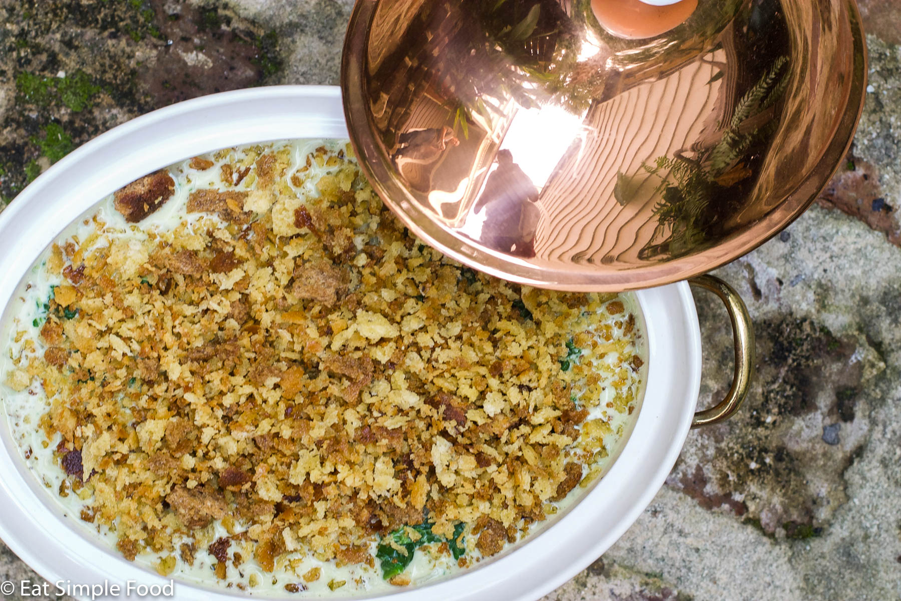 Top view of Spinach Gratin (uncooked) with a Crunchy breaded topping in a copper baking pan on a brick floor.