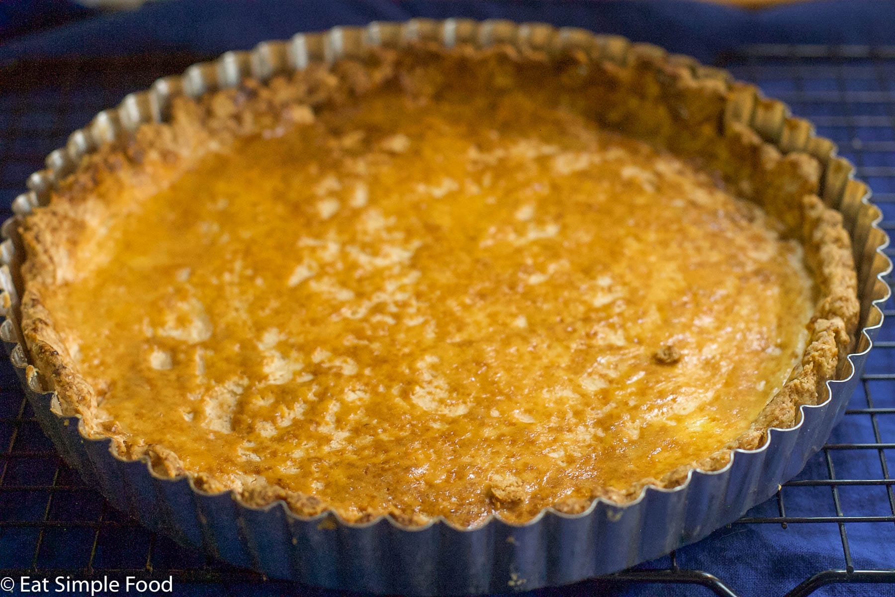 Side view of golden tart pastry cooked in a silver tart shell on an elevated wire rack.